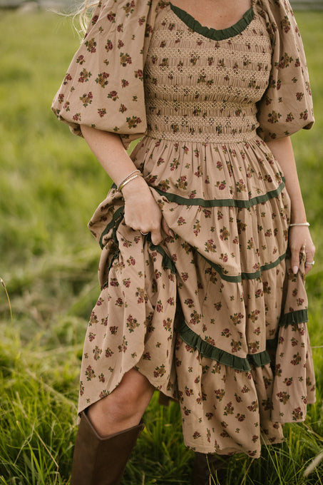 A woman stands in a grassy field wearing the Ivy City Co Roselyn Dress in Desert Blossom, a cotton midi with brown floral print, puffed sleeves, and green trim. She holds one side of her dress and wears brown boots; her face is not visible.