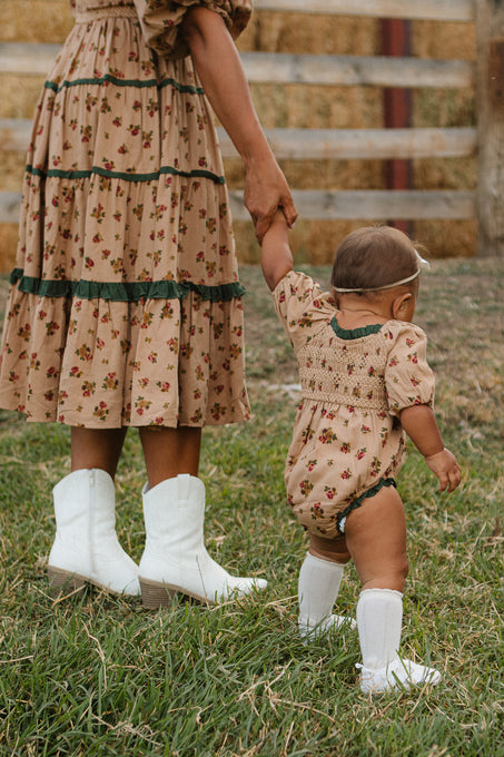 A woman and baby hold hands on the grass in matching floral outfits. The baby wears Ivy City Co’s Baby Roselyn Romper in Desert Blossom with tall white socks and a headband. A wooden fence is visible in the background.