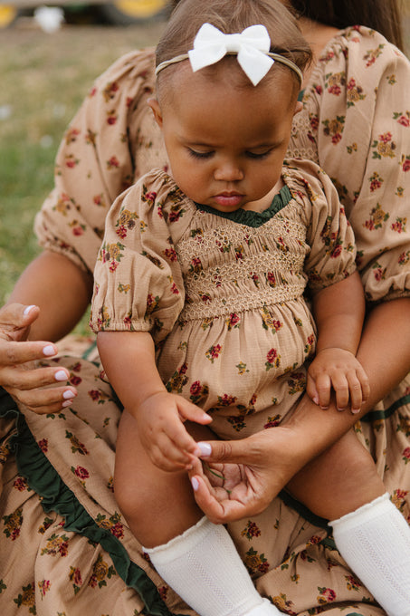 A baby in the Ivy City Co Baby Roselyn Romper in Desert Blossom, white knee-high socks, and a white bow headband sits on a woman’s lap outdoors. The woman also wears a matching floral dress.