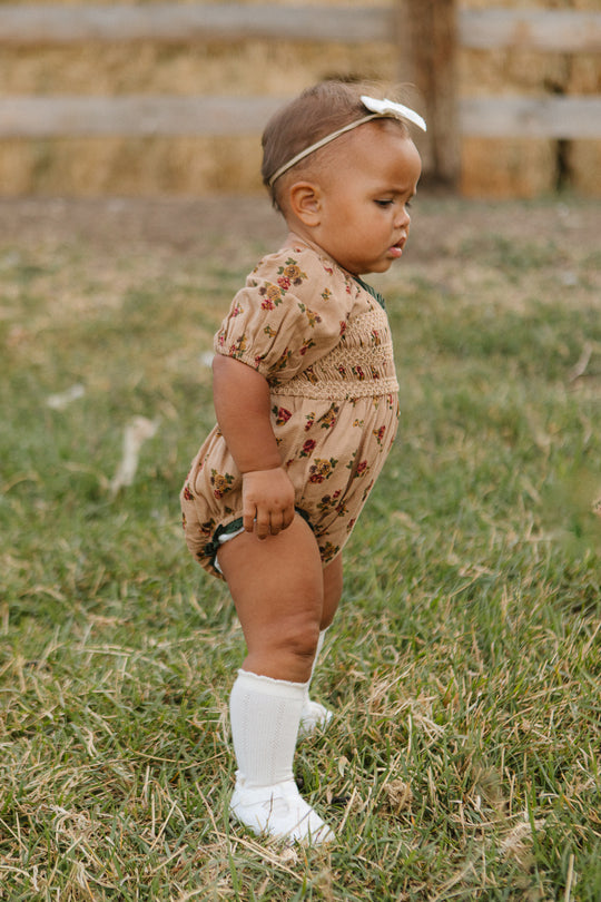 A toddler in the Ivy City Co Baby Roselyn Romper in Desert Blossom, white knee-high socks, and a white headband stands on grass with a wooden fence and hay bales in the background.