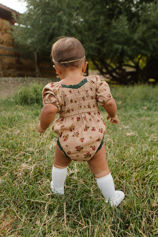 A baby wearing the Ivy City Co Baby Roselyn Romper in Desert Blossom, white knee-high socks, and a headband stands and walks on outdoor grass, facing away from the camera with trees and a wooden fence behind.