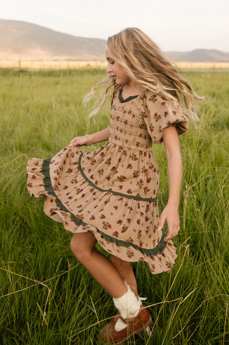 A young girl twirls in a grassy field, wearing the Ivy City Co Mini Roselyn Dress in Desert Blossom—a beige floral midi dress with puffed sleeves—paired with brown shoes and white socks, hills and sky creating a charming backdrop.