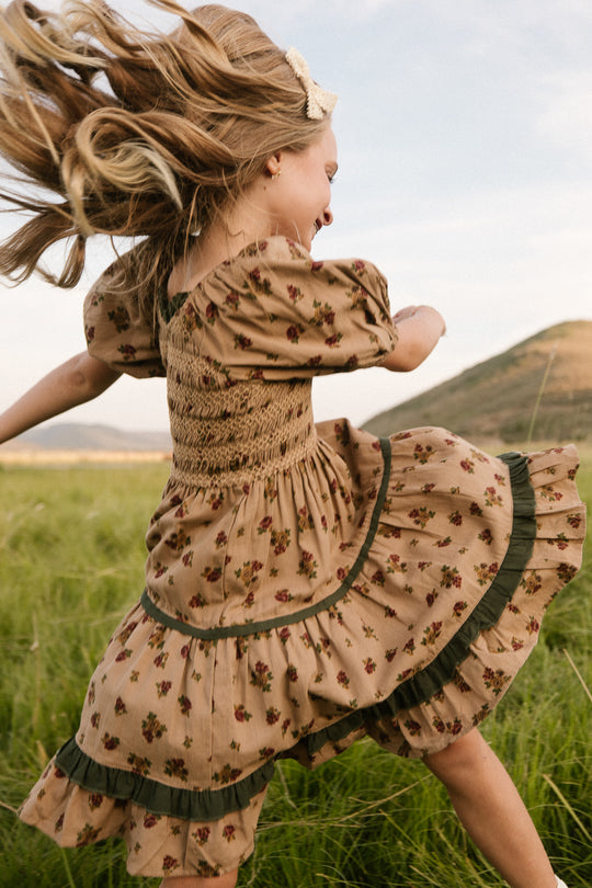 A young girl twirls in a grassy field, wearing the Ivy City Co Mini Roselyn Dress in Desert Blossom—an airy girls midi dress with puff sleeves and a ruffled hem. Her blonde hair flies as she spins, with a green hill behind her.