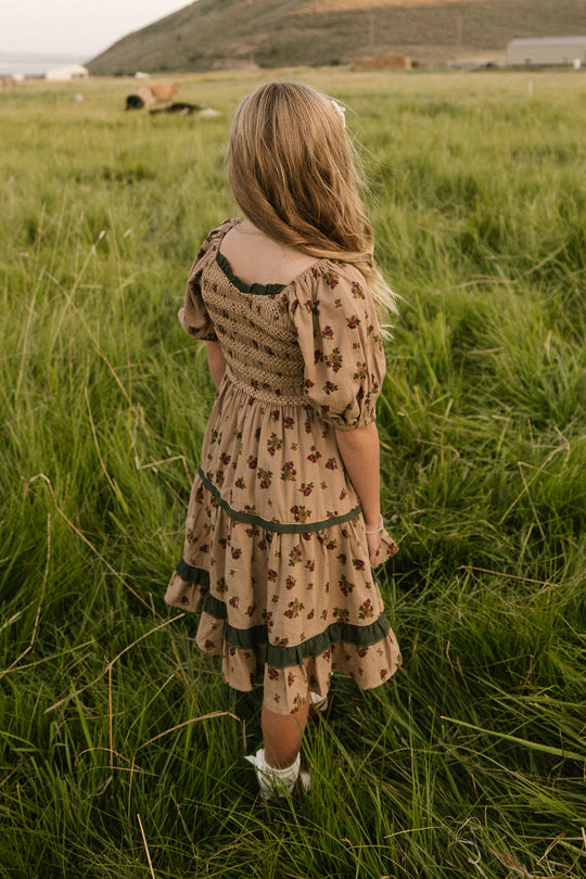 A young girl with long blonde hair, wearing the Mini Roselyn Dress in Desert Blossom by Ivy City Co, stands in a grassy field facing away, with rolling hills and a distant cow highlighting the tranquil rural scene.