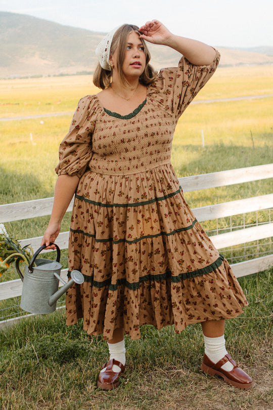 A woman in the Ivy City Co Roselyn Dress in Desert Blossom stands on grass by a wooden fence, holding a watering can with flowers. She gazes into the distance, wearing a white headscarf as a scenic field stretches behind her.