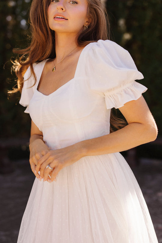 A young woman with long, light brown hair stands outdoors in the sunlight, wearing the Ivy City Co Ballerina Dress in White—featuring puff sleeves and a square neckline—with greenery softly blurred in the background.