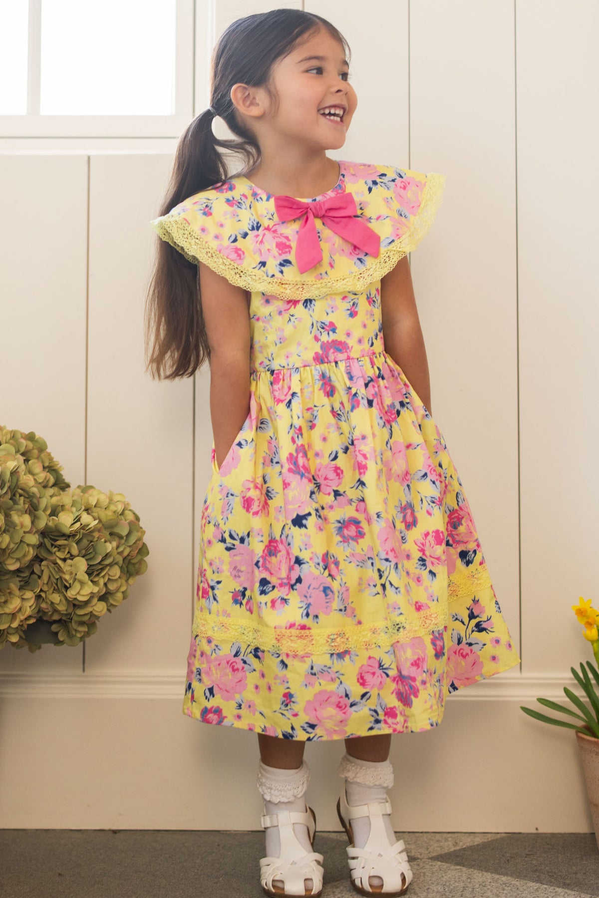 A young girl with long dark hair smiles indoors in the Ivy City Co Mini Aster Dress in As You Dream Floral, featuring a lace-trimmed collar and pink bow at the neck, paired with white sandals. Flowers and a window are behind her.