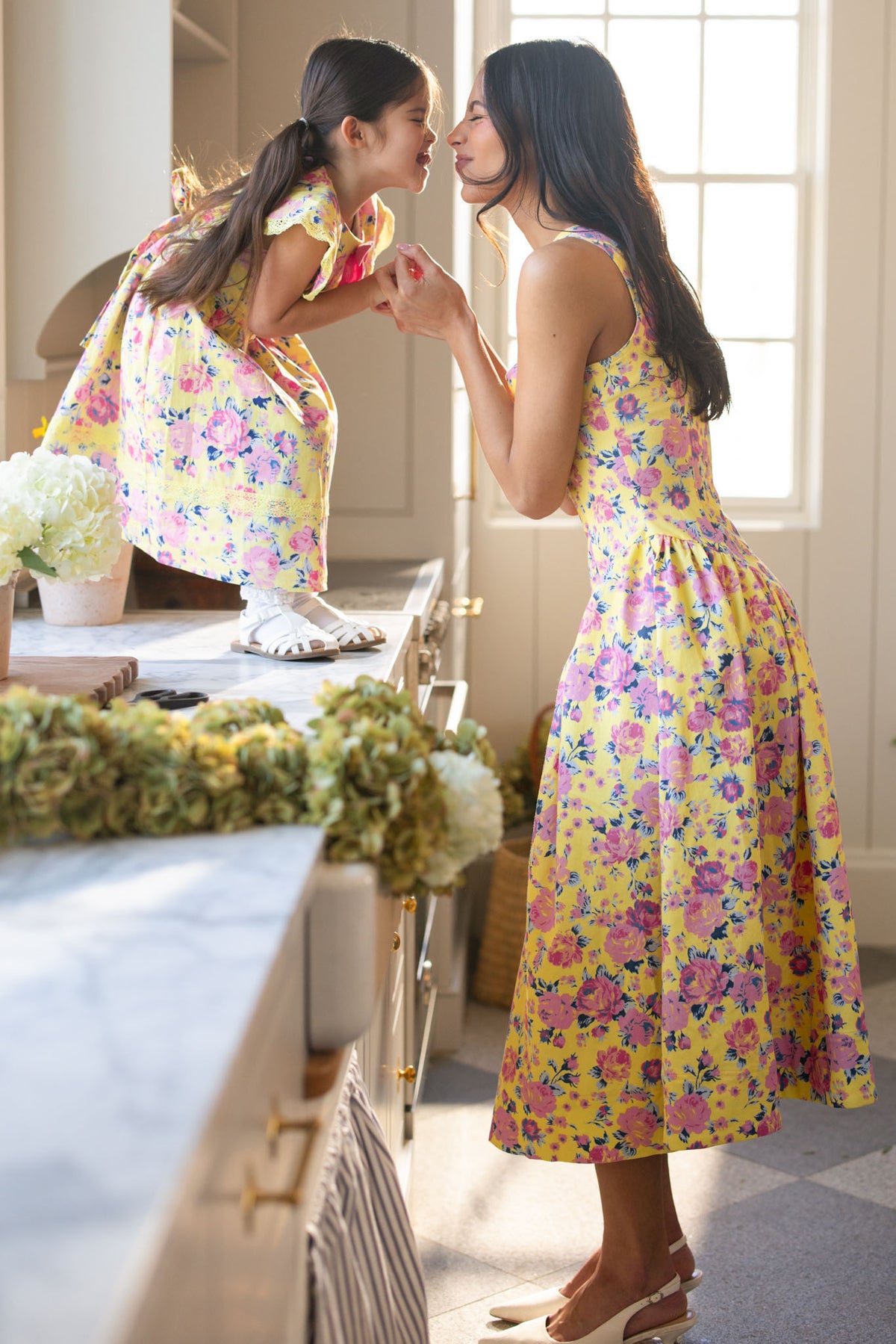 A woman and young girl smile and touch noses in a bright kitchen, wearing matching Ivy City Co Edith Dresses in As You Dream Floral. The girl stands on the counter as the woman stands beside her, holding her hands.