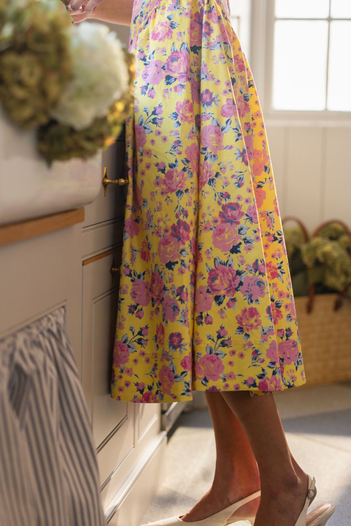 A person wears the Ivy City Co Edith Dress in As You Dream Floral, standing barefoot in a sunlit kitchen with flowers on the counter and a woven basket filled with greenery in the background.