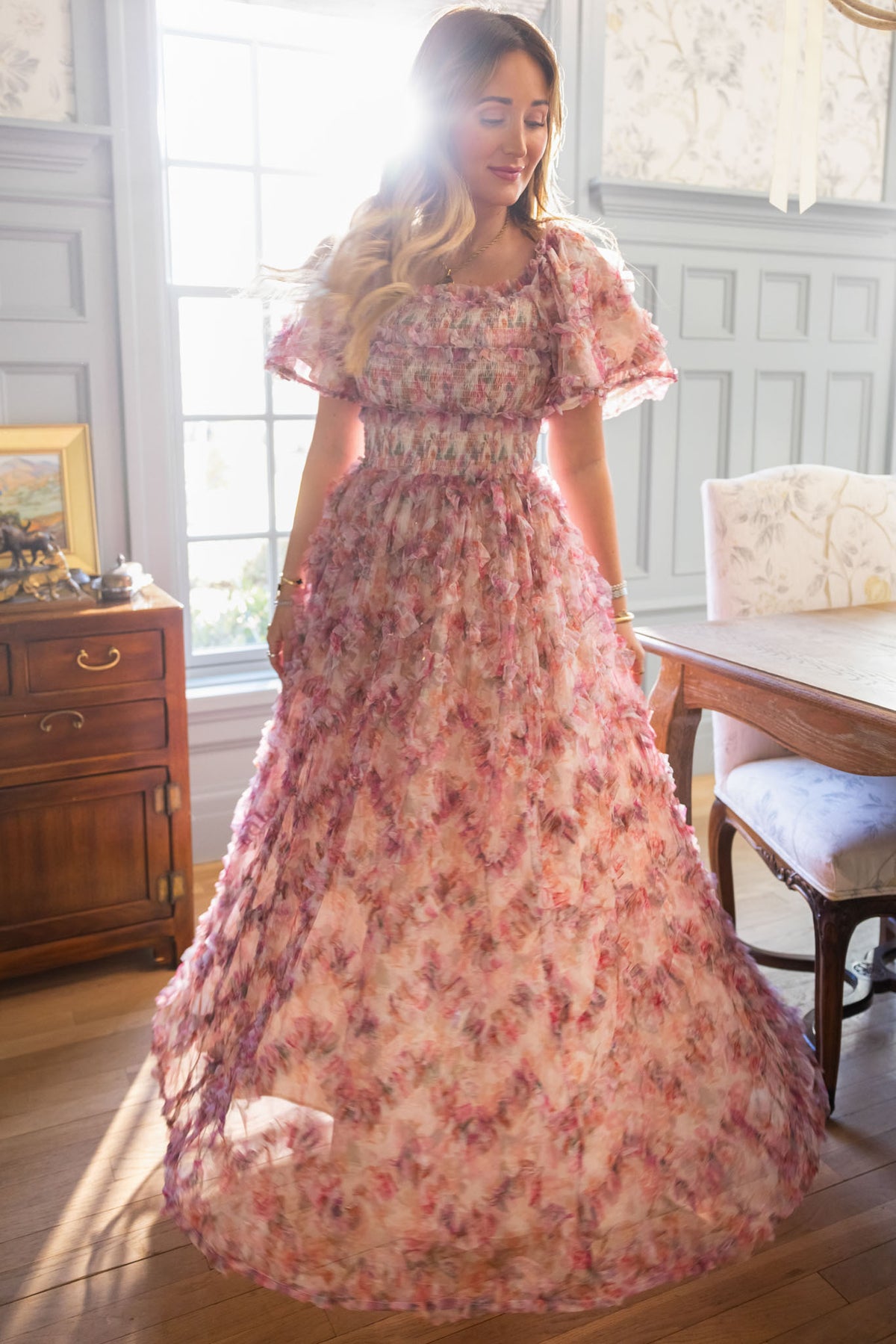 A woman in the Ivy City Co Florentina Dress in Alluring Floral—a pink floral design exuding timeless femininity—stands in a sunlit room with wooden furniture and pale gray paneled walls, soft sunlight glowing behind her.