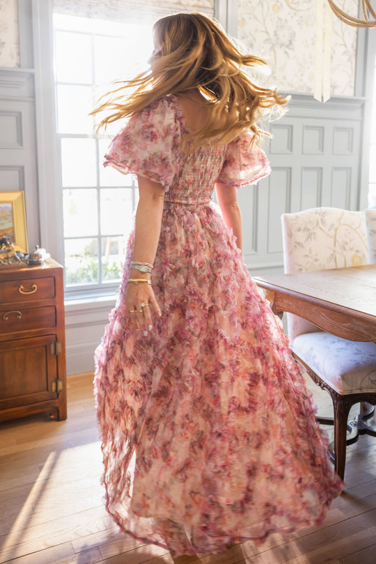 A woman twirls in a sunlit room wearing the Ivy City Co Florentina Dress in Alluring Floral, its timeless femininity enhanced by pink blooms as sunlight floods through large windows onto gray paneled walls and wooden floors.
