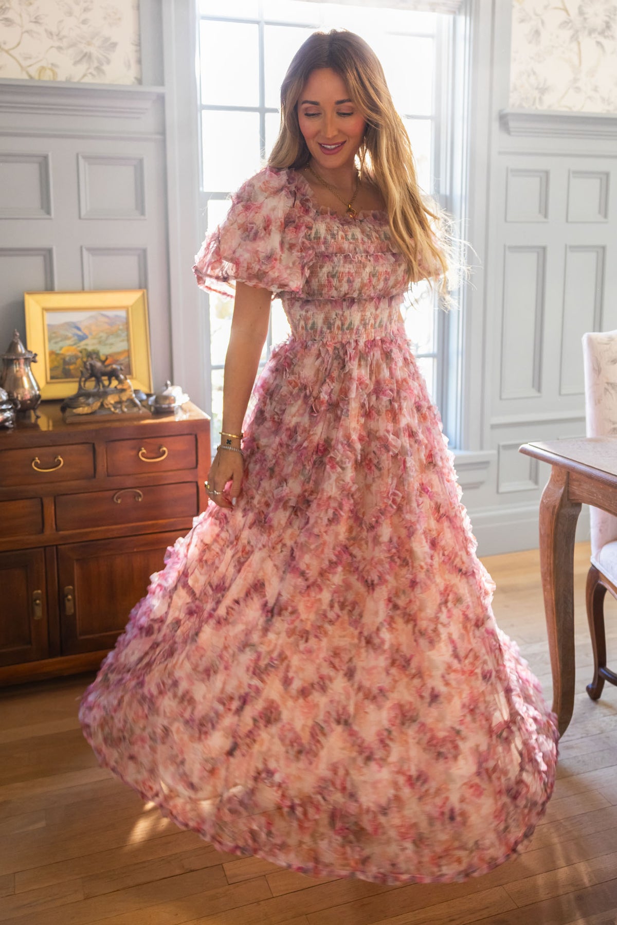 A woman twirls in a sunlit room, radiating timeless femininity in Ivy City Co’s Florentina Dress in Alluring Floral, surrounded by gray paneled walls, wooden floors, and a brown sideboard with framed art and silver accents.