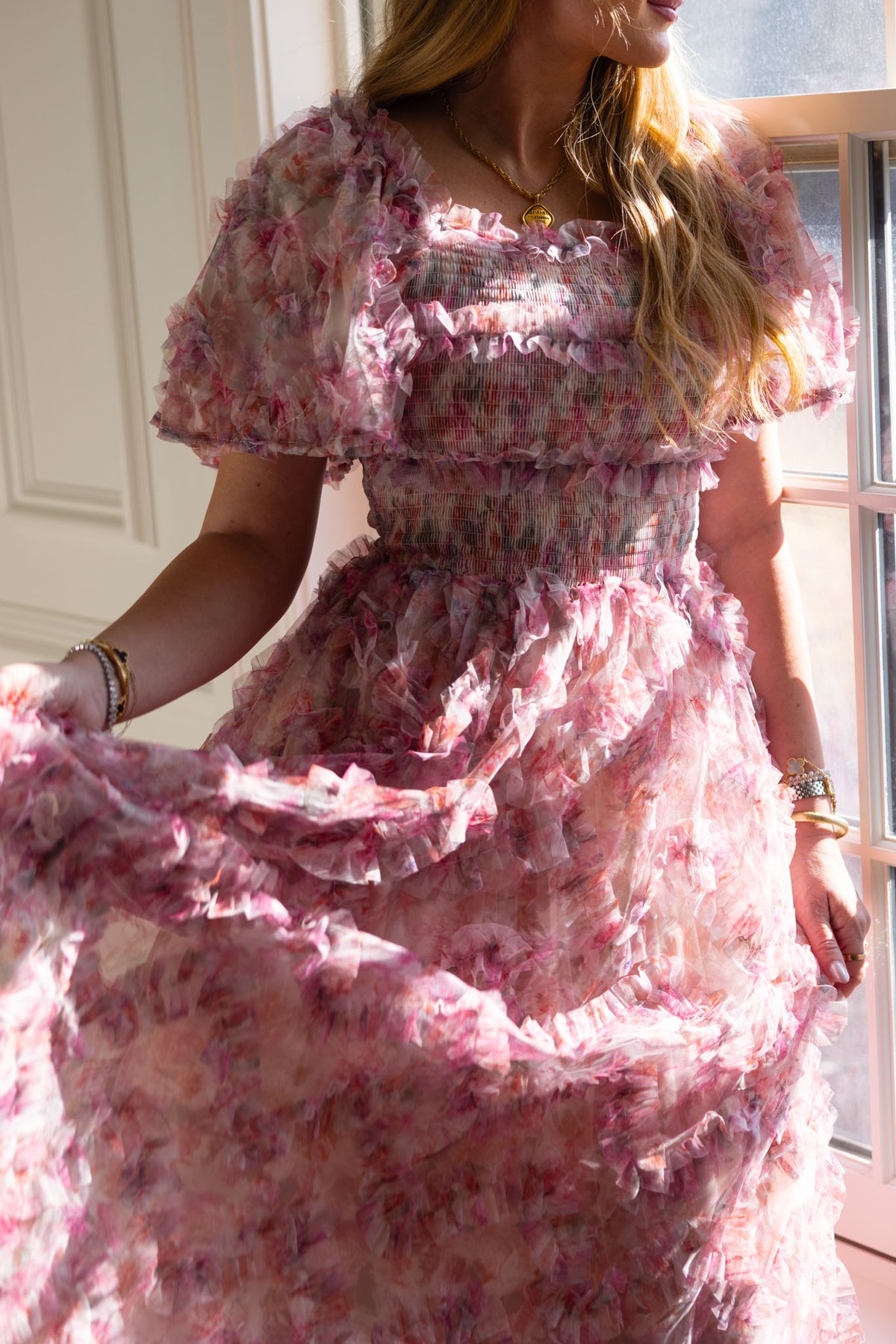 A woman in sunlight wears Ivy City Co’s Florentina Dress in Alluring Floral—its pink ruffled print and puff sleeves evoke timeless femininity. She shows off its flowy skirt, her wavy hair loose and gold jewelry catching the light.