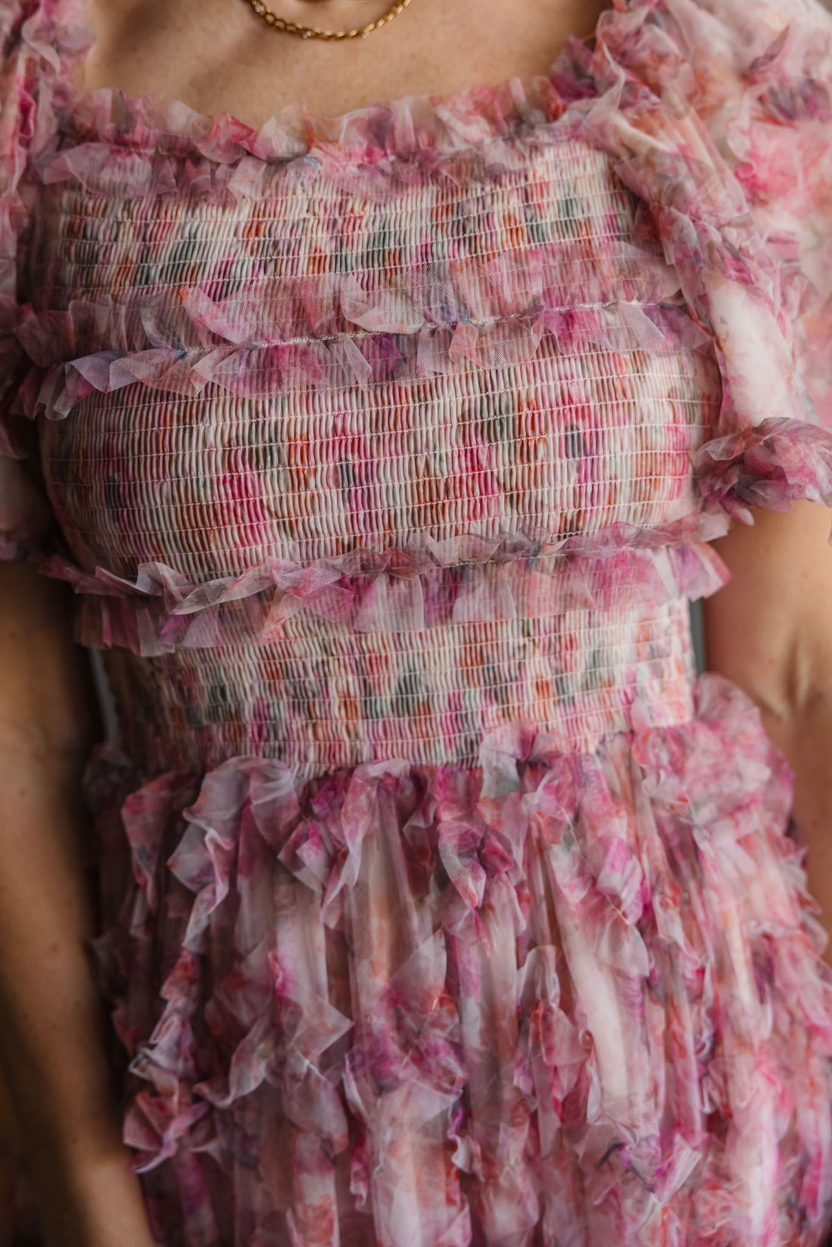 A close-up of a person wearing the Ivy City Co Florentina Dress in Alluring Floral—a pink, ruffled piece with a smocked bodice, short sleeves, and sheer fabric—styled with a gold chain necklace for timeless femininity.