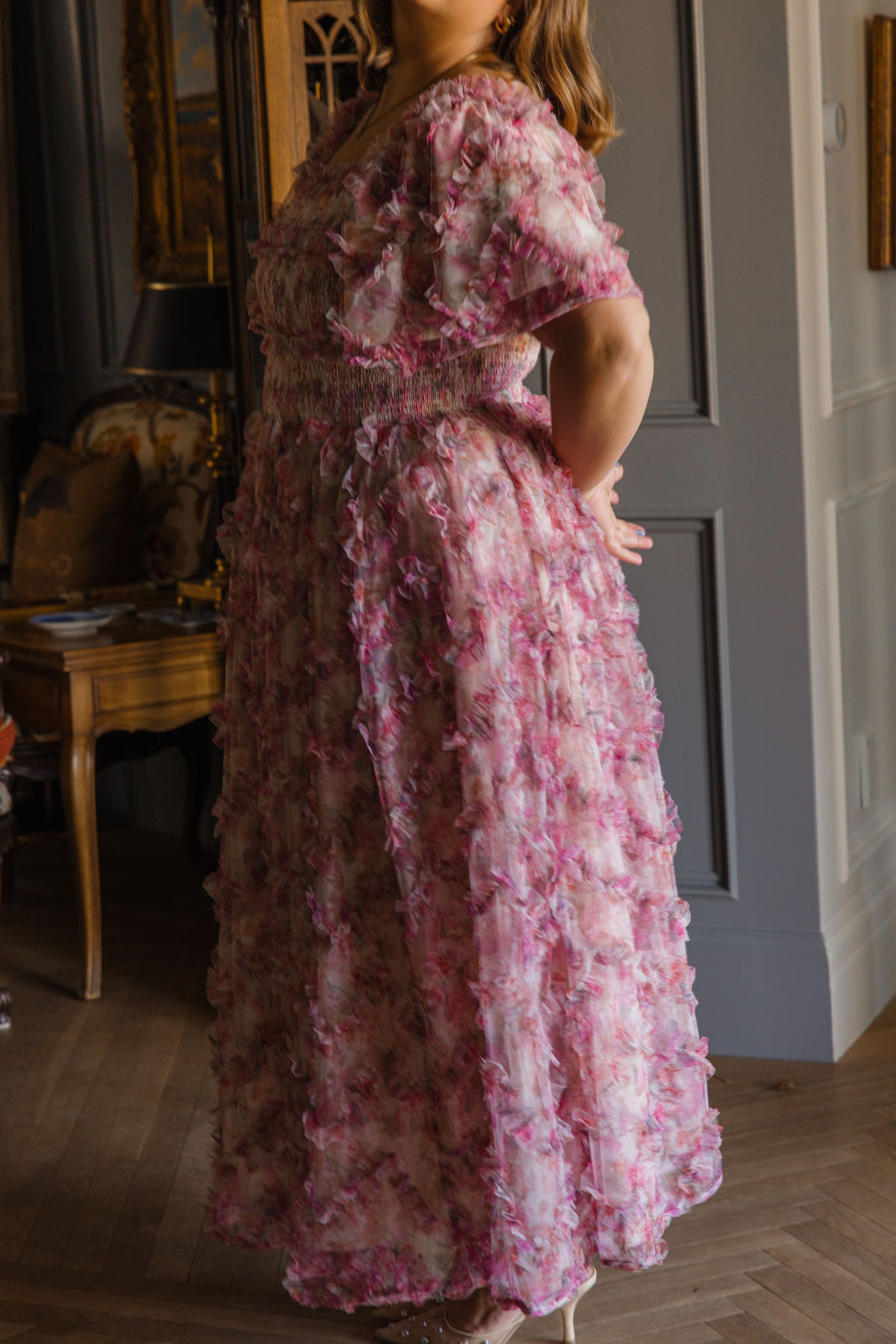 A woman in the Florentina Dress in Alluring Floral by Ivy City Co stands indoors, hands behind her back. Soft lighting and classic decor enhance the flowy pink floral print, highlighting her timeless feminine style.