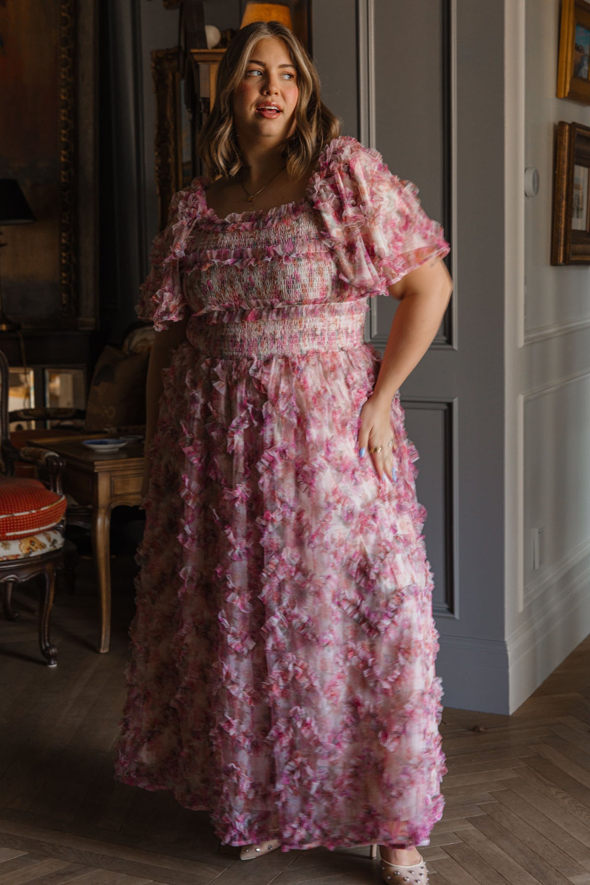 A woman with shoulder-length brown hair smiles softly indoors, wearing the Ivy City Co Florentina Dress in Alluring Floral—a pink and purple textured dress with short puff sleeves—amid classic decor and wooden furniture.