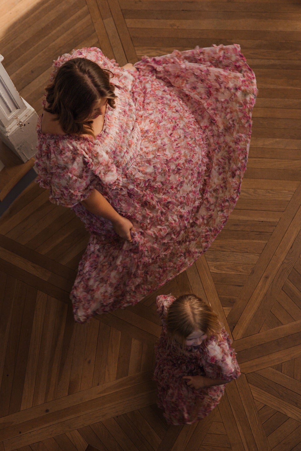 A woman and a young girl wear matching Ivy City Co Florentina Dress in Alluring Floral; the woman twirls, her dress fanned out on the wooden floor, while the girl gazes up in admiration, capturing a timeless moment.