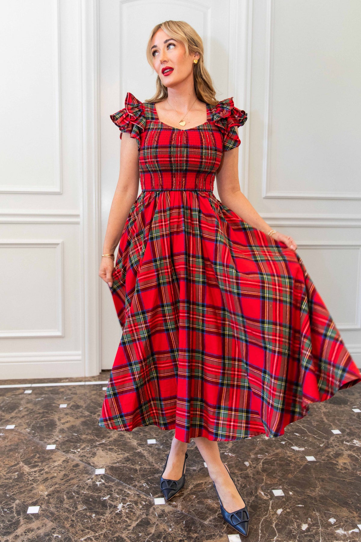 Woman wearing a red plaid dress standing against a white paneled wall.