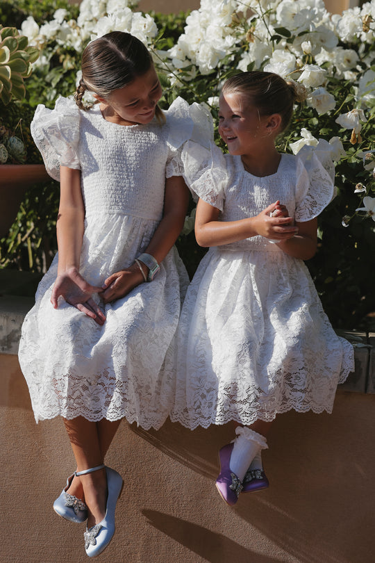 Two girls in Ivy City Co’s Mini Hattie Dress in White Lace sit together, smiling with white flowers behind them. One wears blue shoes and the other purple, making this sweet moment in matching dresses picture-perfect.