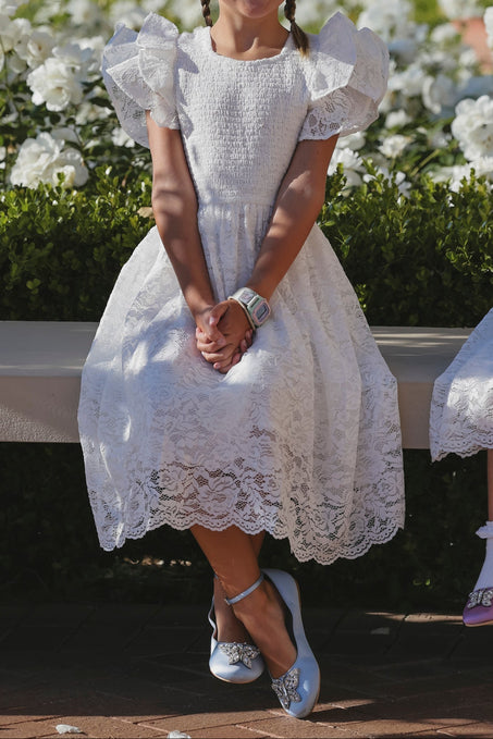 A young girl sits on a bench in the Mini Hattie Dress in White Lace by Ivy City Co, wearing blue shoes with bows and a watch. Her hands are folded in her lap as white flowers bloom in the background.