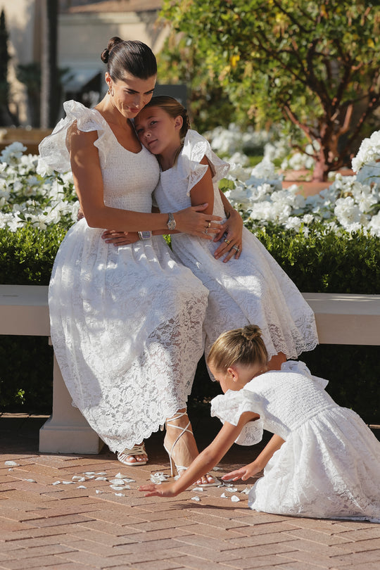 A woman in a white lace dress hugs a young girl wearing the Mini Hattie Dress in White Lace by Ivy City Co, while another girl in a matching midi dress picks up flower petals. White flowers and greenery complete the beautiful backdrop.