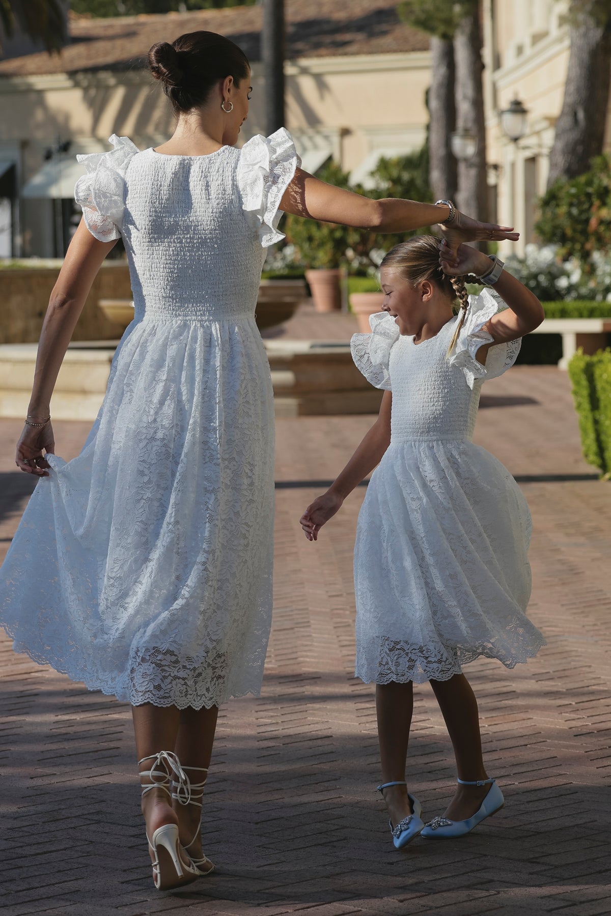 A woman and young girl dance outdoors in matching Ivy City Co Hattie Dresses in White Lace, holding hands as they twirl along a path with greenery, their romantic dresses fluttering in the sunny breeze.