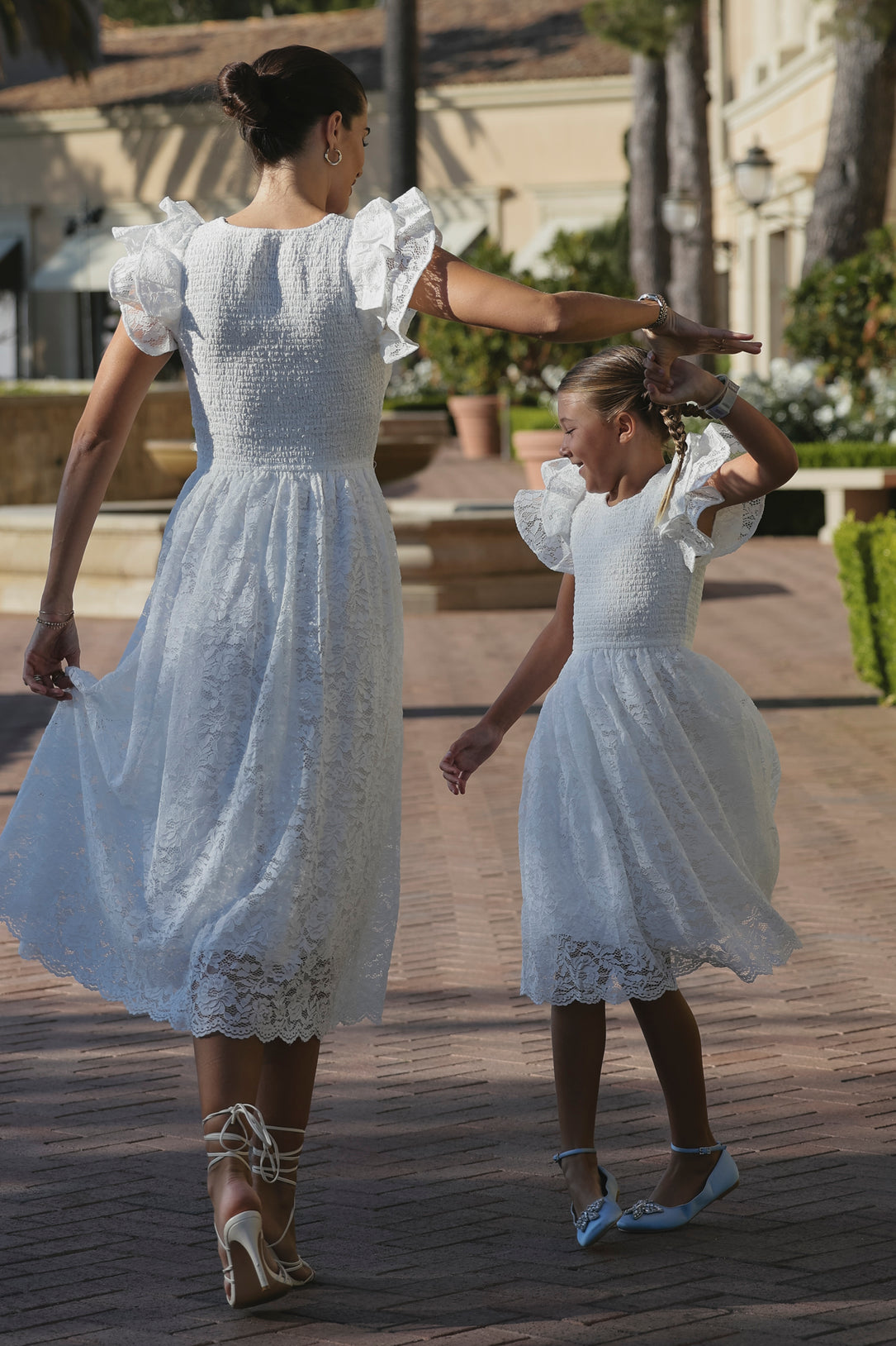 A woman and girl joyfully spin together on a sunlit brick path, both wearing Ivy City Co’s Mini Hattie Dress in White Lace and dress shoes, surrounded by greenery.