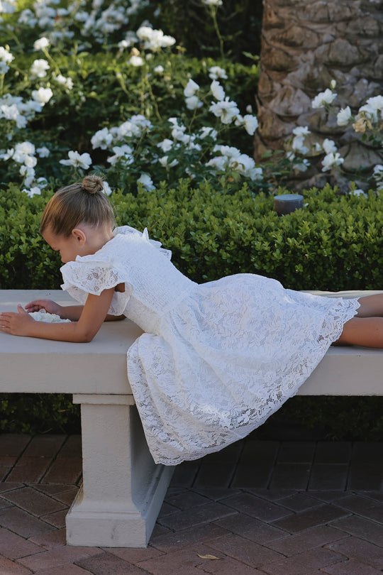 A young girl wearing the Ivy City Co Mini Hattie Dress in White Lace lies on her stomach on a stone bench, gazing at something in her hands, surrounded by blooming white flowers and lush green bushes in a garden.
