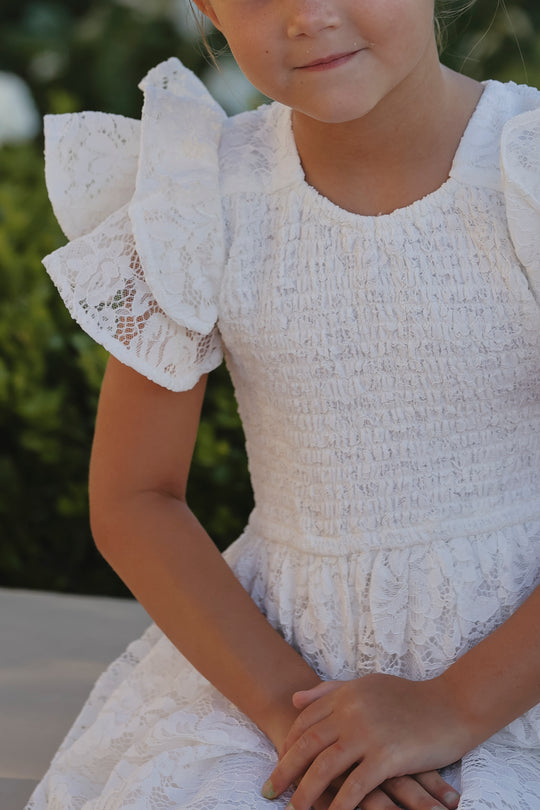 A young girl in the Ivy City Co Mini Hattie Dress in White Lace with puffed sleeves sits with her hands folded in her lap against a blurred green backdrop, her face partially visible as she looks slightly downward.
