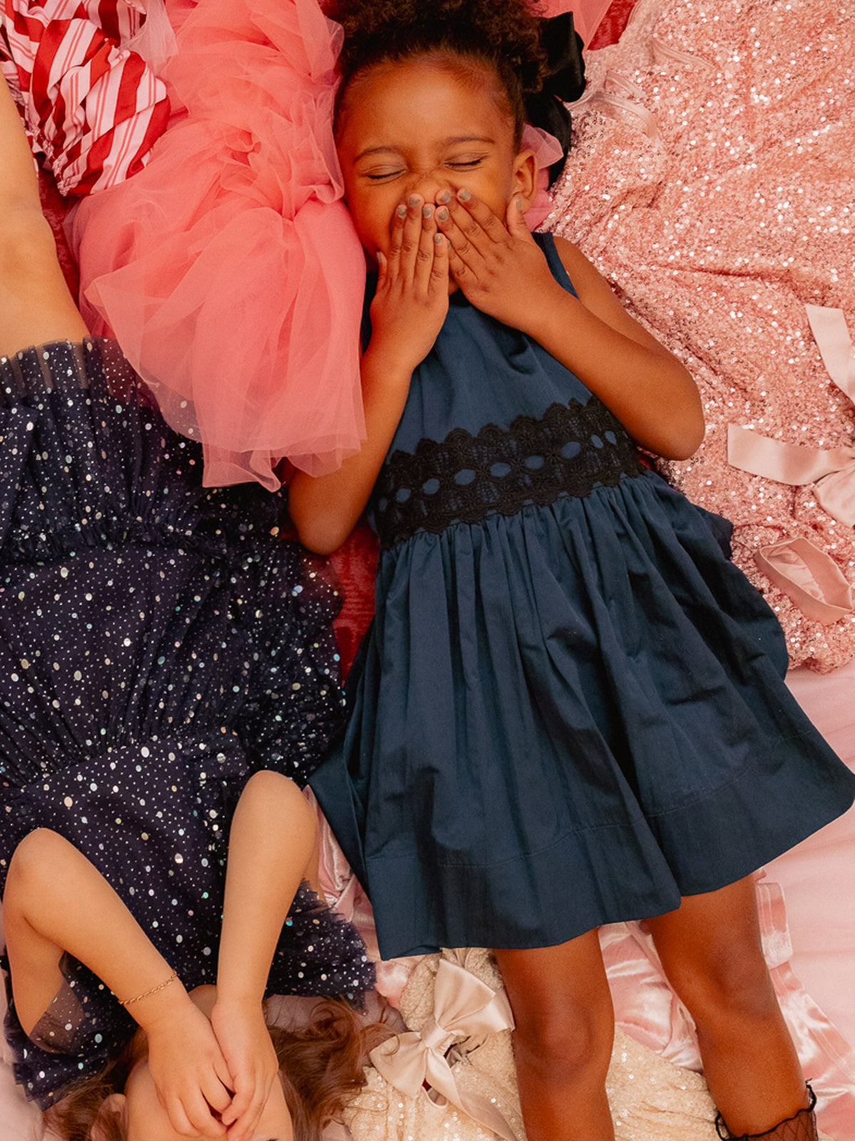 Two young children in sparkly dresses lie on a pink, glittery surface, smiling and covering their mouths with their hands. One wears a navy sleeveless dress with a delicate lace waistband; the other is dressed in the charming Ivy City Co Mini Greyson Dress in Navy with white dots.
