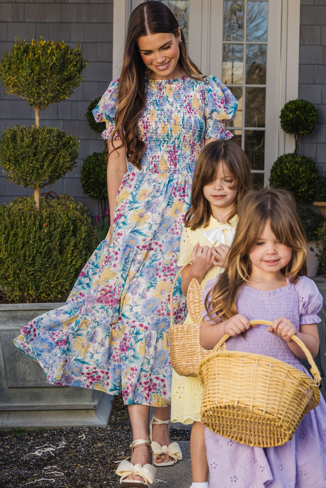 A woman in the Ivy City Co Cordelia Dress in Pretty Plumeria smiles outdoors with two young girls holding wicker baskets, enjoying a sunny garden day—perfect for showcasing their favorite spring dresses.