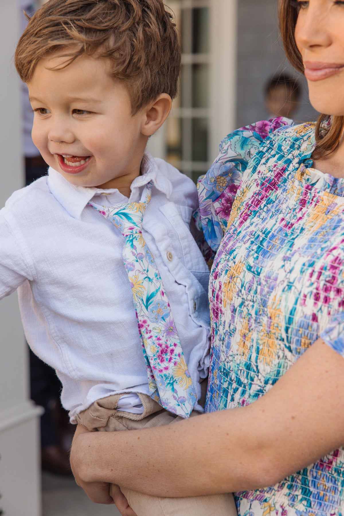 A young boy in a white shirt and Ivy City Co's Ties in Pretty Plumeria smiles while being held by a woman in a multicolored textured dress, outdoors near large windows, ready for a cheerful spring celebration.