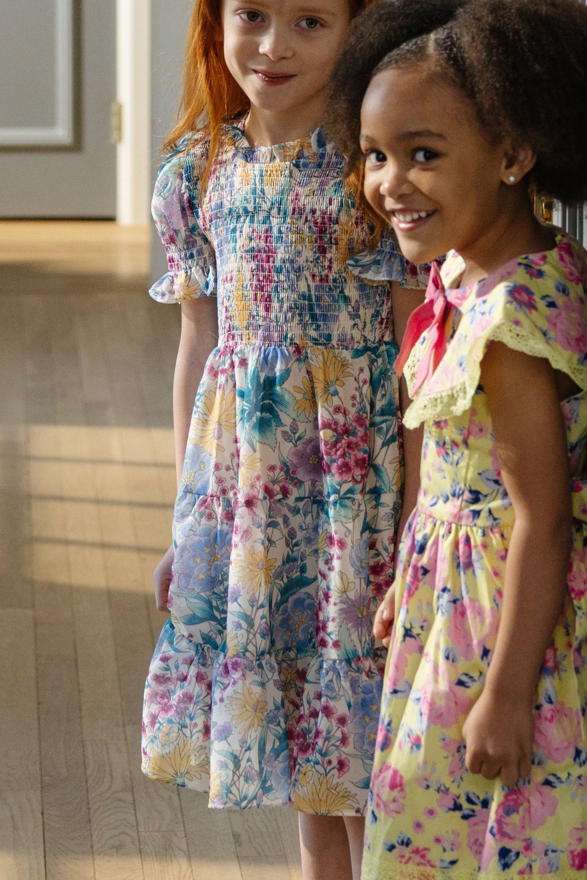 Two young girls stand indoors, smiling during spring celebrations. One wears a yellow floral dress; the other dons the Ivy City Co Mini Cordelia Dress in Pretty Plumeria. Sunlight shines on the wooden floor, capturing joyful family moments.