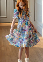 A young girl with long red hair wears the Ivy City Co Mini Cordelia Dress in Pretty Plumeria—a floral dress with short sleeves, pleats, and ruffles—standing indoors on a wooden floor, holding out the skirt and looking down. Perfect for spring celebrations.