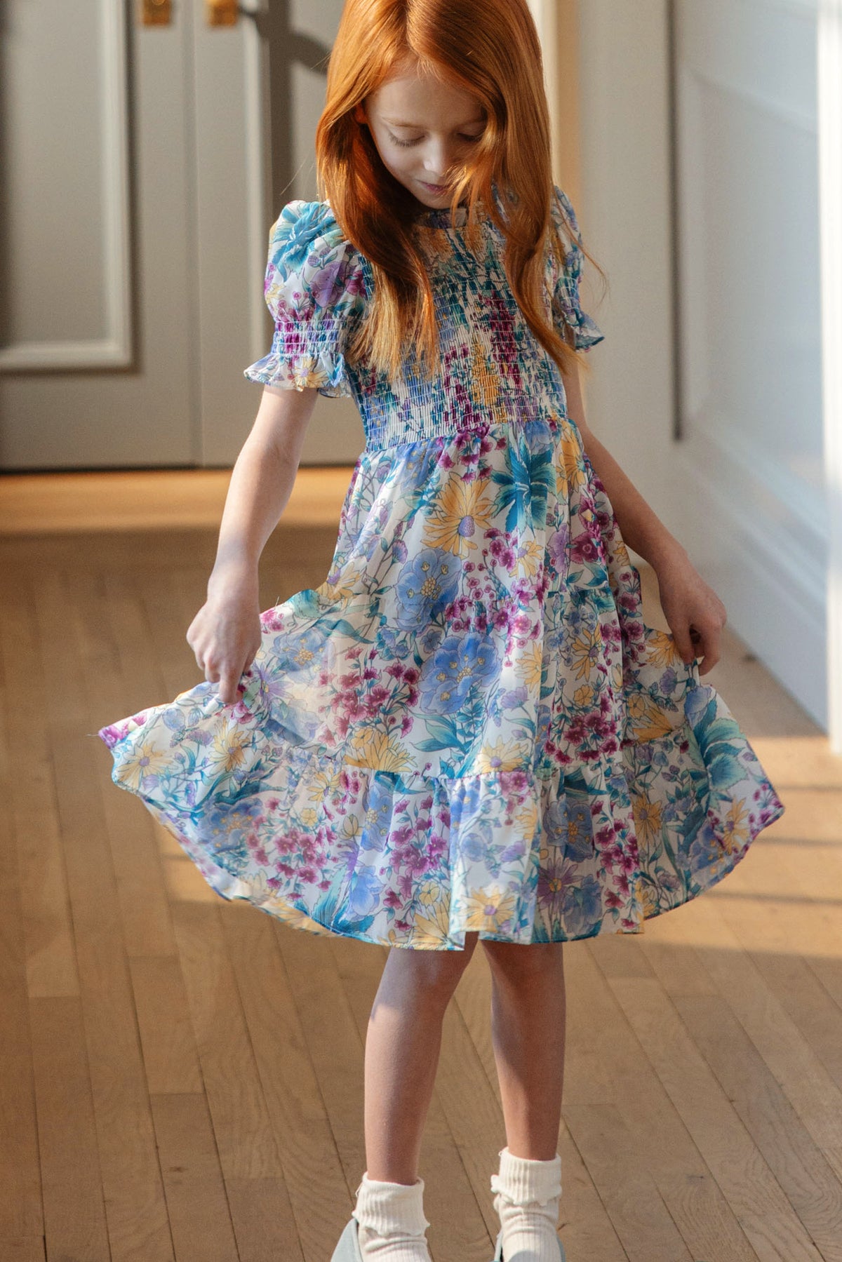 A young girl with long red hair wears the Ivy City Co Mini Cordelia Dress in Pretty Plumeria—a floral dress with short sleeves, pleats, and ruffles—standing indoors on a wooden floor, holding out the skirt and looking down. Perfect for spring celebrations.