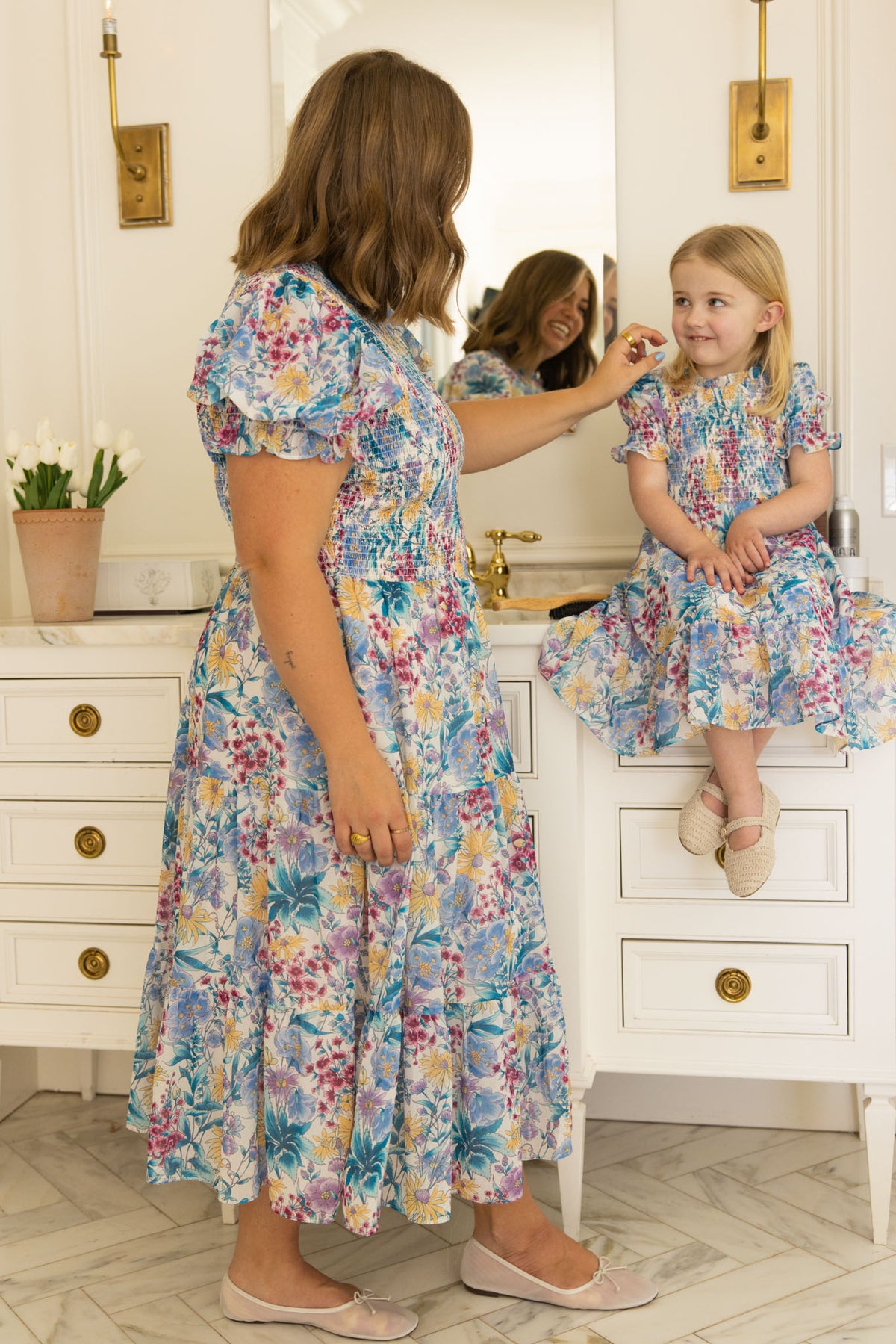 A woman and a young girl share smiles in a bright bathroom, capturing sweet family moments in matching Ivy City Co Mini Cordelia Dresses in Pretty Plumeria—the girl perched on the counter while the woman gently touches her face.