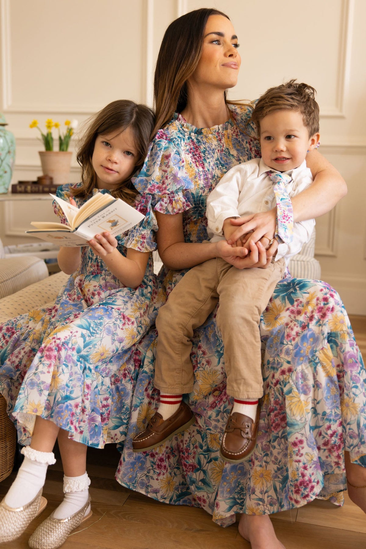 A woman wearing the Ivy City Co Mini Cordelia Dress in Pretty Plumeria sits indoors with a young girl reading a book beside her and a young boy in khakis and a tie on her lap, capturing joyful family moments. All three appear happy.