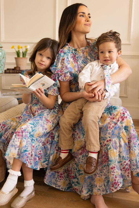 A woman wearing the Ivy City Co Mini Cordelia Dress in Pretty Plumeria sits indoors with a young girl reading a book beside her and a young boy in khakis and a tie on her lap, capturing joyful family moments. All three appear happy.