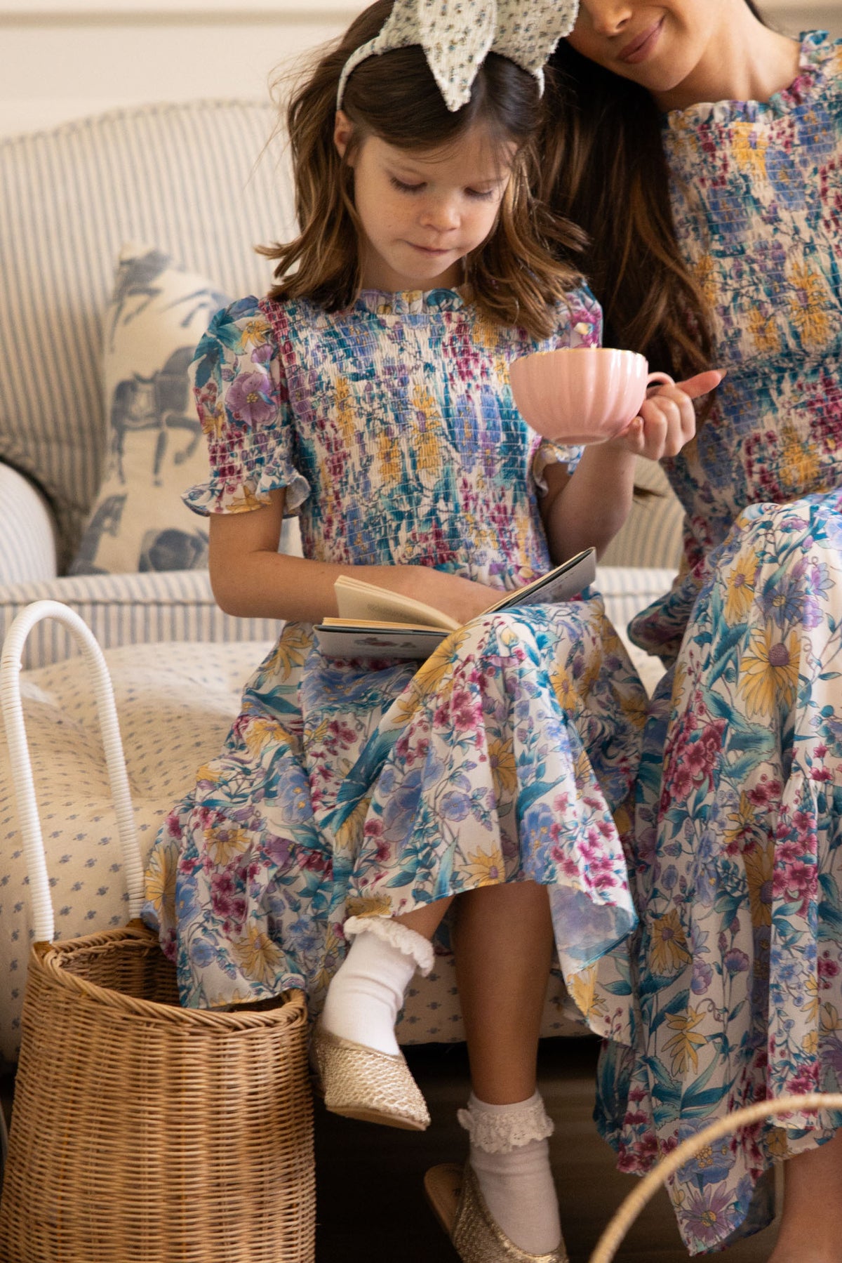 A young girl in the Ivy City Co Mini Cordelia Dress in Pretty Plumeria sits on a couch, reading a book and holding a pink teacup beside a woman in a matching dress. A woven basket rests at her feet, capturing sweet family moments.
