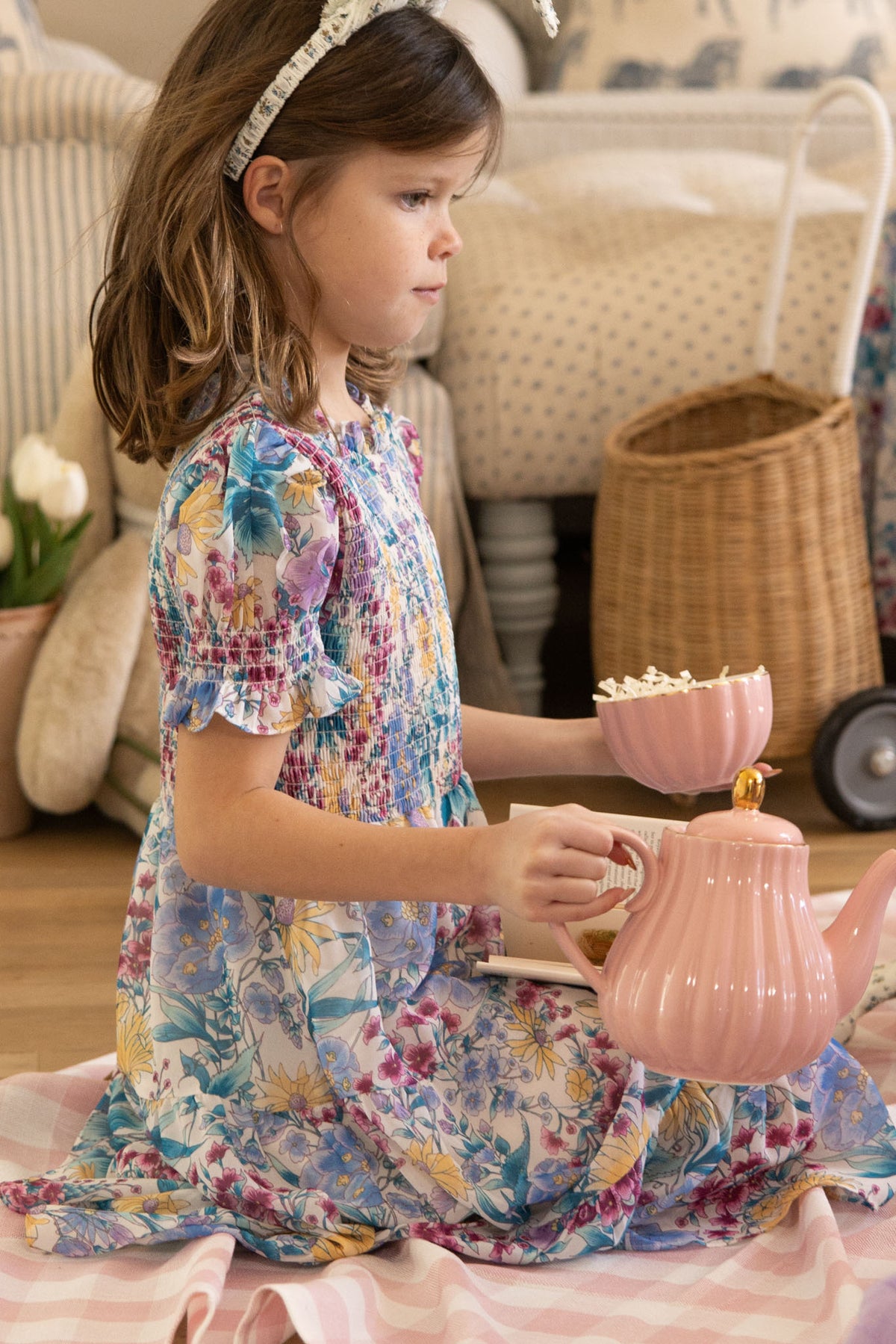 A young girl in the Ivy City Co Mini Cordelia Dress in Pretty Plumeria sits on a checkered blanket, holding a pink teapot and popcorn-filled teacup—ready for a charming indoor tea party or spring family celebration.