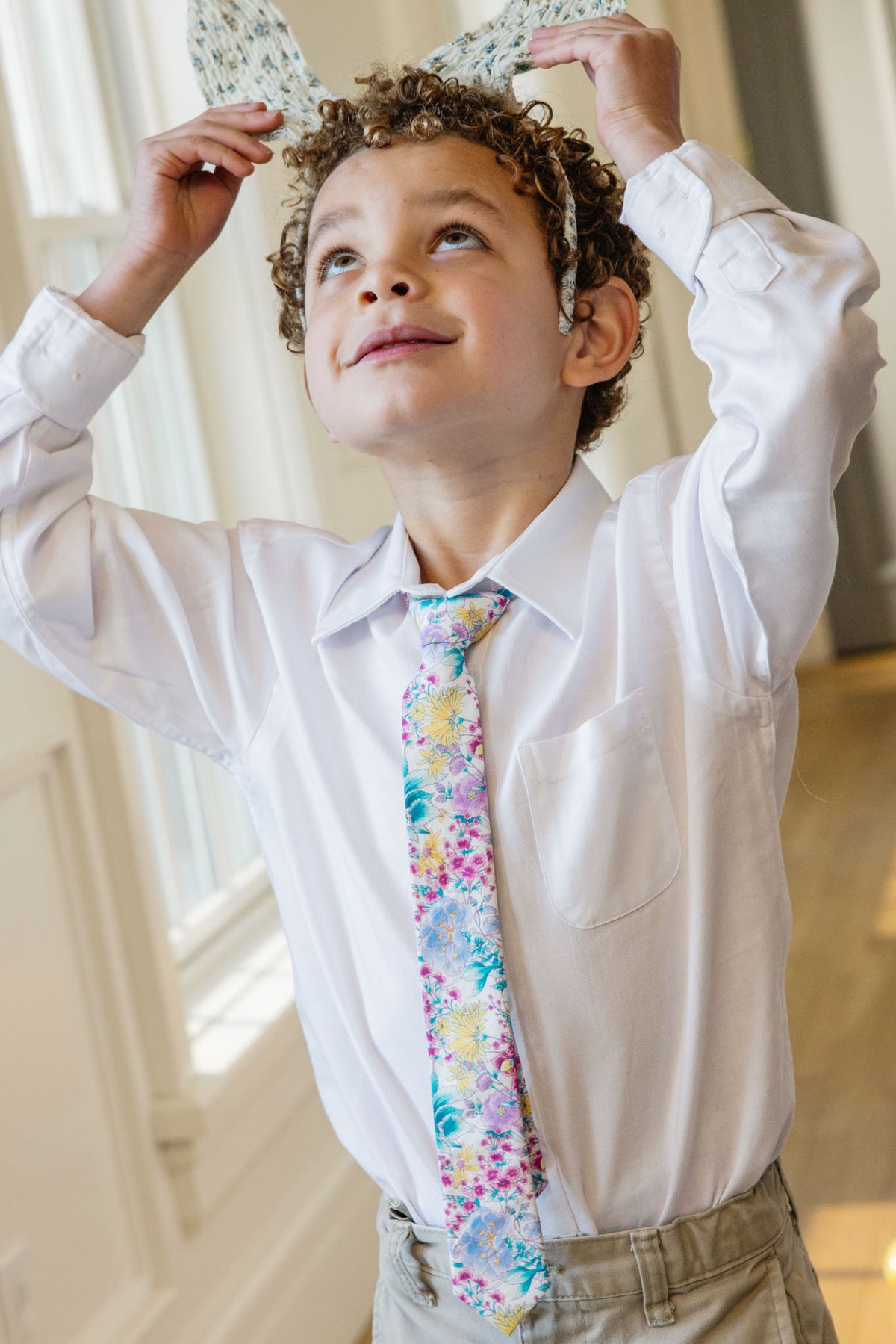 A young child in a white dress shirt, khaki pants, and the Ivy City Co Ties in Pretty Plumeria holds bunny ears above their head in a bright room, ready for festive spring fun.