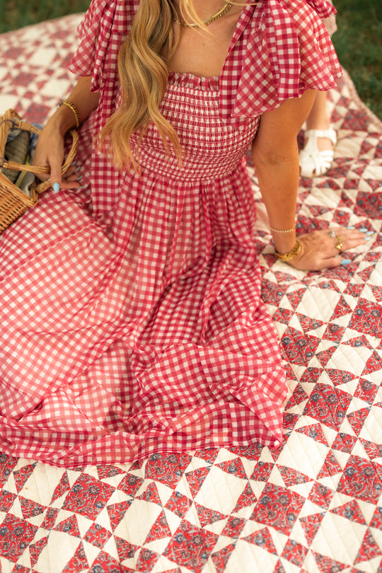 A woman wears the Ivy City Co Rayna Dress in Red Gingham, sitting outdoors on a patterned quilt with a wicker basket. Her wavy hair falls over her shoulder and she accessorizes with gold jewelry; only her upper body and legs are visible.