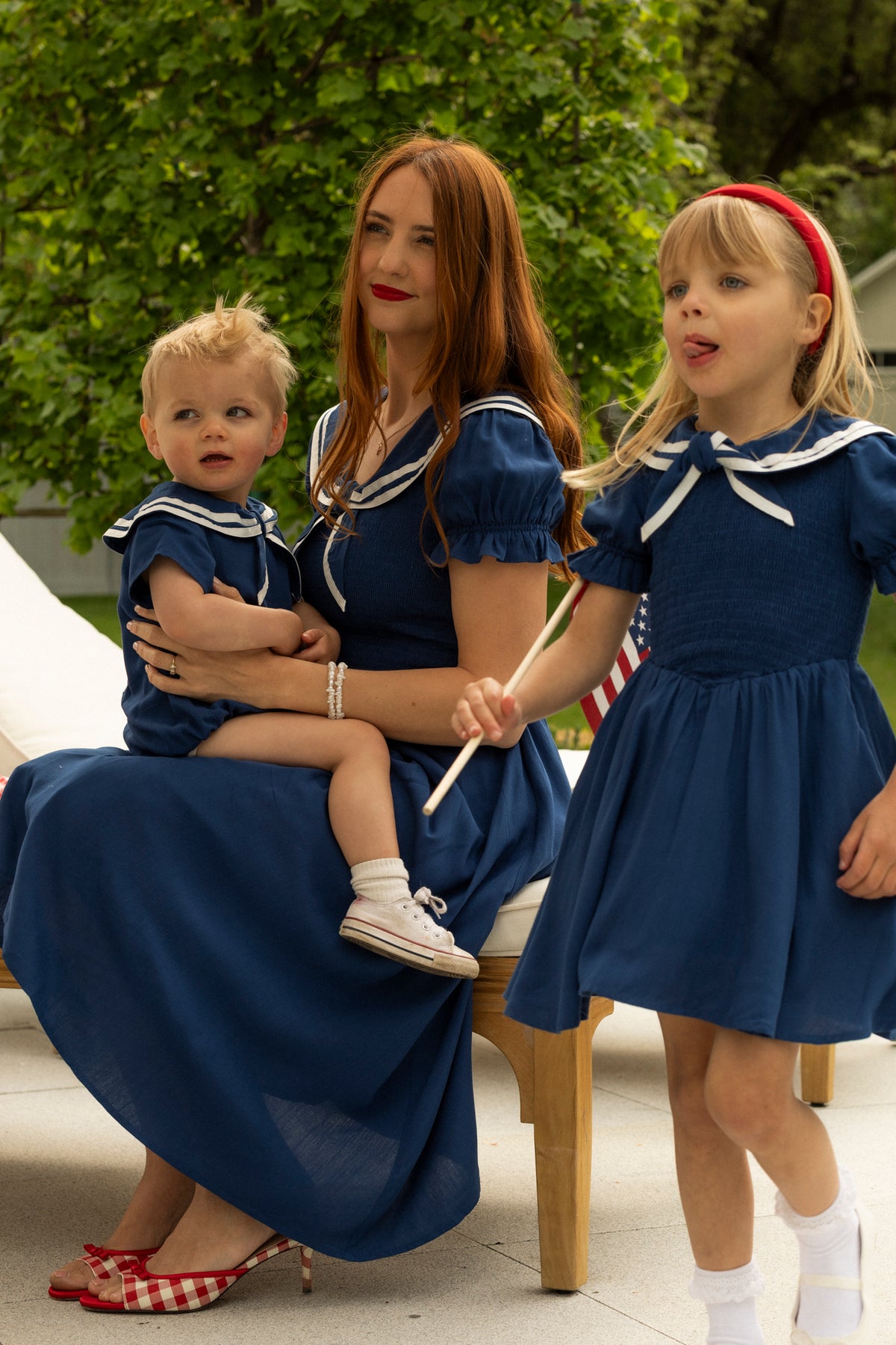 A woman with long red hair sits on a bench holding a child, while an older girl waves an American flag nearby. All three wear the Ivy City Co Alice Dress in Sailor Navy with white collars, enjoying time outdoors by greenery.