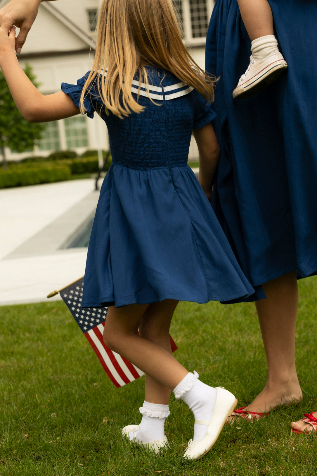 A young girl in the Ivy City Co Mini Alice Dress in Navy holds hands with an adult carrying a child. She waves an American flag on the grass near a house, creating a classic matching Mom and Me moment.