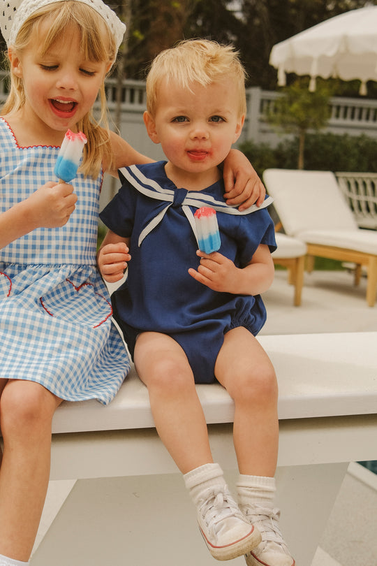 Two young children sit by a pool, smiling and eating red, white, and blue popsicles. One wears a blue gingham dress; the other is in the Ivy City Co Baby Sailor Romper in Navy with white sneakers for the perfect nautical look.