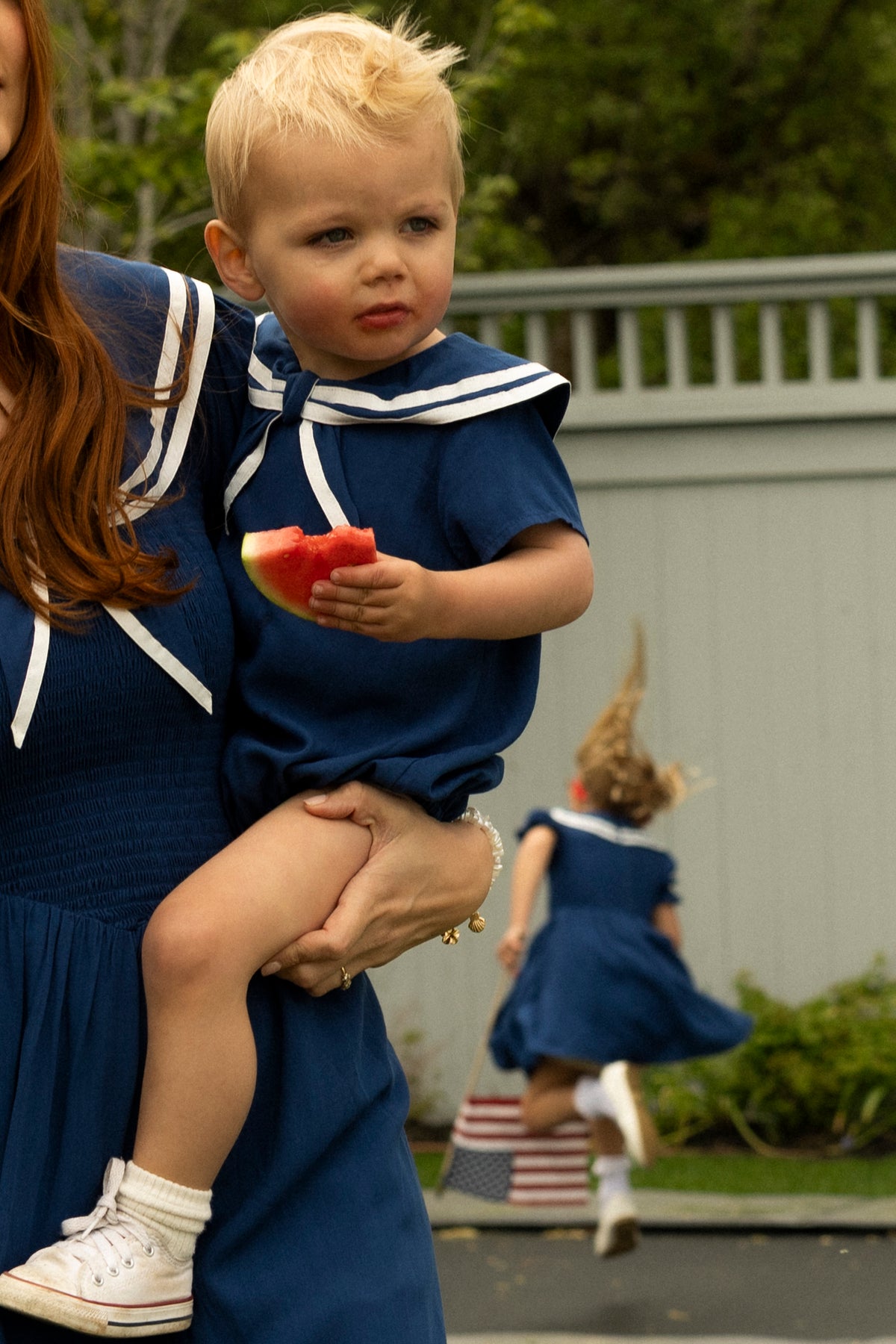 A small child in the Ivy City Co Baby Sailor Romper in Navy eats watermelon while held by an adult. In the background, another child in a matching romper runs toward greenery.