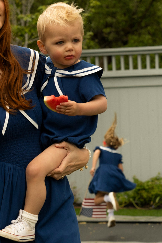 A small child in the Ivy City Co Baby Sailor Romper in Navy eats watermelon while held by an adult. In the background, another child in a matching romper runs toward greenery.