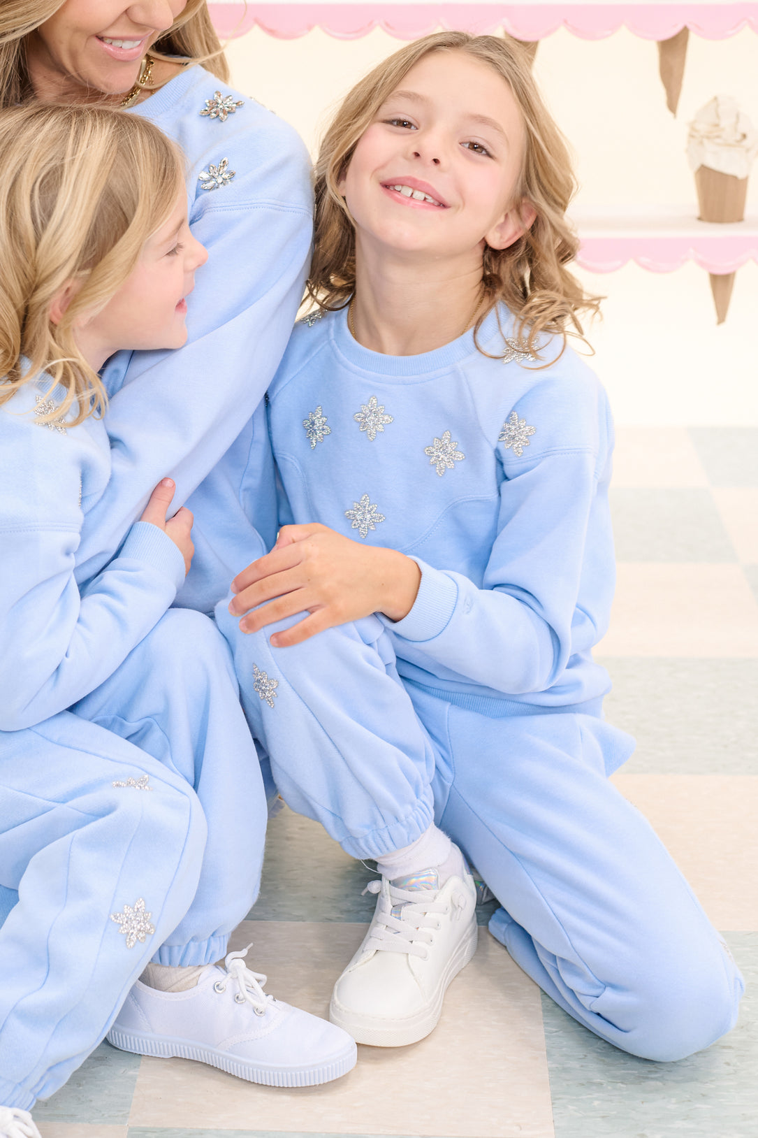 Two children wear Ivy City Co's Mini Casey Jogger Sweatpants in Light Blue with snowflake designs, smiling on a checkered floor. A similarly dressed adult is partly visible nearby, with pastel ice cream cone decor in the background.