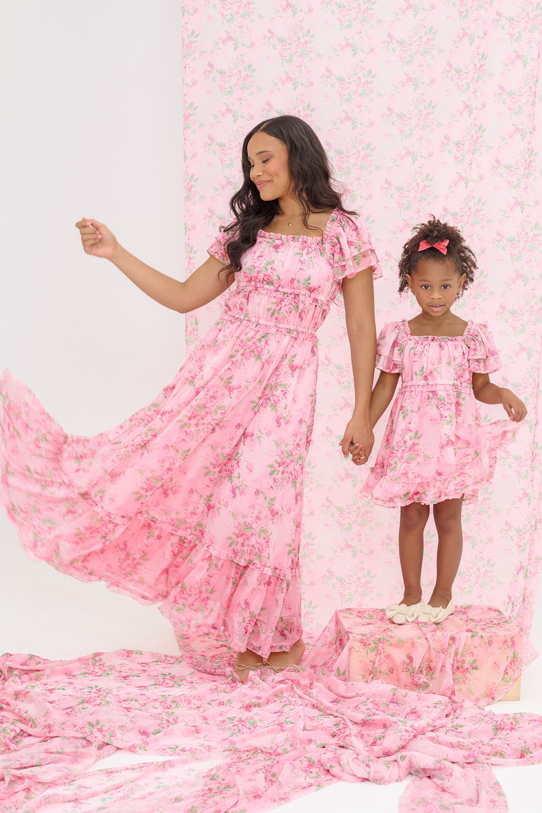 A woman and young girl wear matching Ivy City Co Juliana Dresses in Rose Trance. The nursing-friendly design features pink floral patterns that echo the coordinating backdrop, as they twirl hand-in-hand, with the girl standing on a draped box.