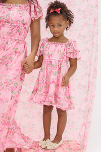A young girl wears the Ivy City Co Mini Juliana Dress in Rose Trance, a pink floral dress with ruffled sleeves and a matching bow, standing hand-in-hand with an adult in coordinating outfits against a pink floral background.